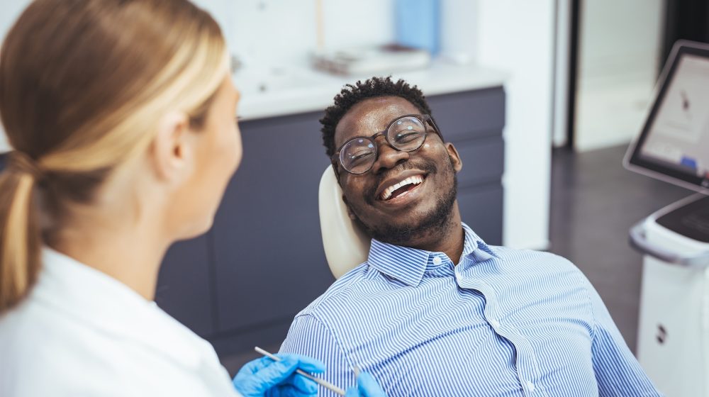 Smiling african guy in dentist chair looking with trust at his doctor, close up. Young African American Man Getting Teeth Treatment With Professional Stomatologist At Modern Clinic